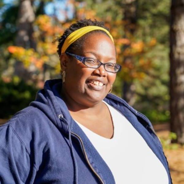 A headshot of Santina. She smiles against a wooded background, wearing a blue sweater and yellow headband.