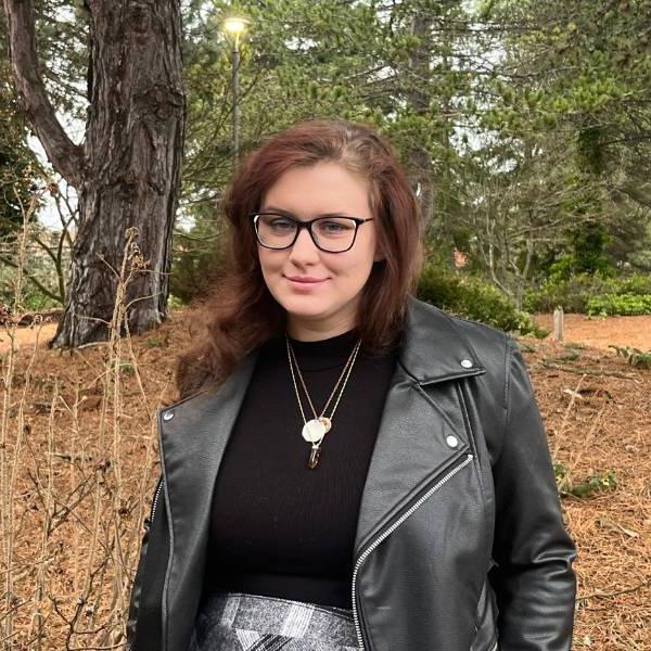 A headshot of Taylor. She smiles against a wooded background, wearing a black shirt and black leather jacket.