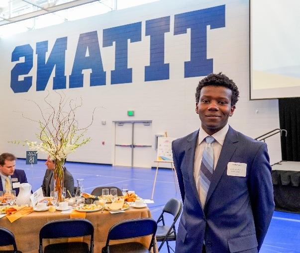 A student in a suit smiles in front of a wall that says "Titan"